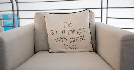Un cuscino chiaro con la scritta “Do small things with great love” appoggiato su una poltrona beige