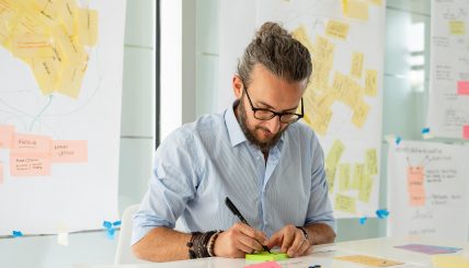 Nicolò scrive su un post-it verde con un pennarello nero. Sullo sfondo, lavagne piene di post-it e schemi rappresentano un processo di brainstorming.