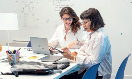 Due designer collaborano a un tavolo con laptop e tablet, sorridendo al lavoro.