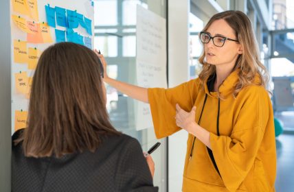 Giulia talks to Ilenia, while they write post it notes and paste them on the wall