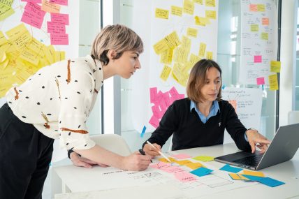 Due designer lavorano insieme con laptop e post-it colorati su un tavolo di progetto.