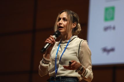 Silvia Riva, holding a microphone and speaking at URCA! 2024