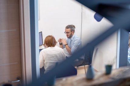 Due persone sedute discutono davanti a uno schermo di computer. Una persona gesticola mentre parla, trasmettendo un senso di dialogo attivo.