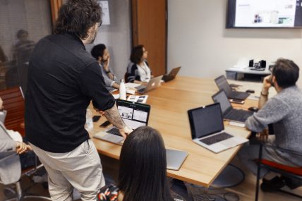 Riunione con più persone attorno a un tavolo, portatili aperti e presentazione proiettata a schermo.