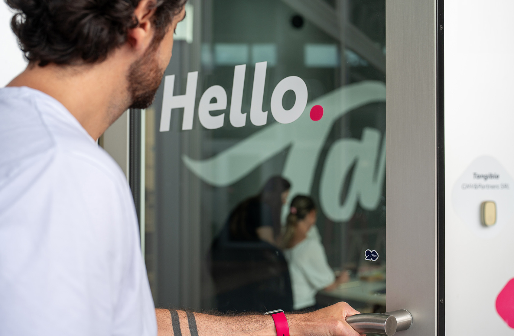 A person opens the door of an office on which "Hello." is written. Through the glass door, two seated people can be seen, intent on their work.