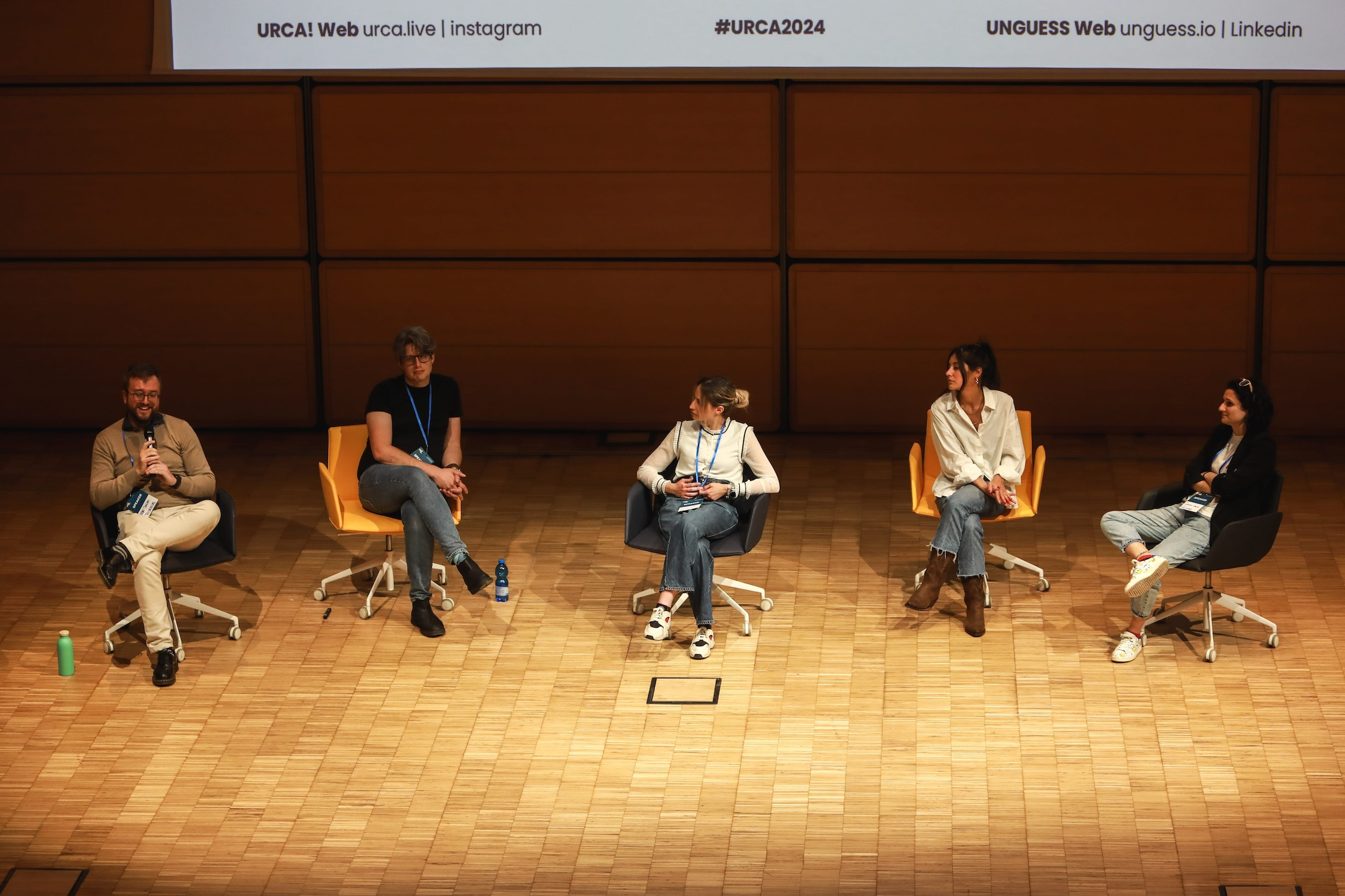 Five speakers sat in colourful chairs lined up on the stage at URCA! 2024 for the panel discussion on the present and future of user research side by side with AI.