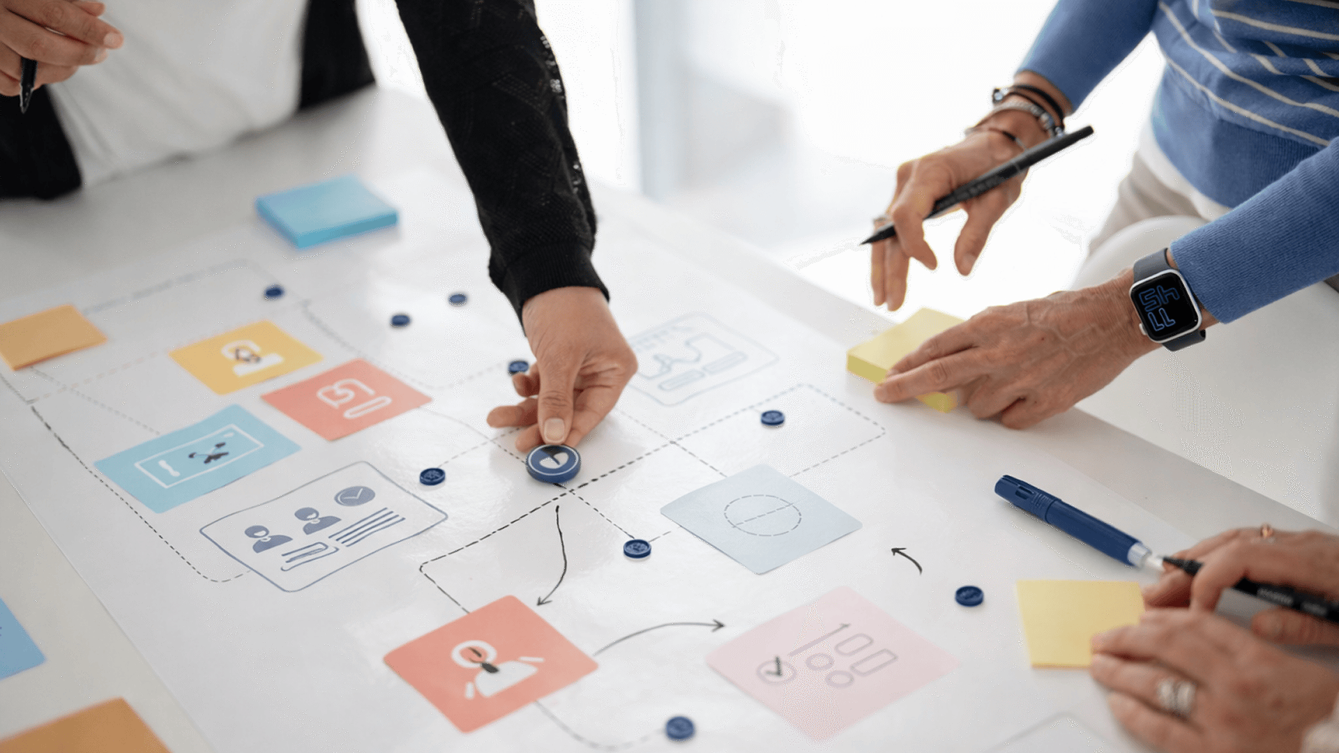 Alcune persone al lavoro su un canvas cartaceo con blocchi colorati, icone e connessioni, mentre posizionano gettoni e prendono appunti.