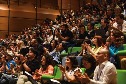 Un'ampia platea di persone sedute su sedie verdi in un auditorium, che applaudono durante una presentazione ad URCA! 2024.