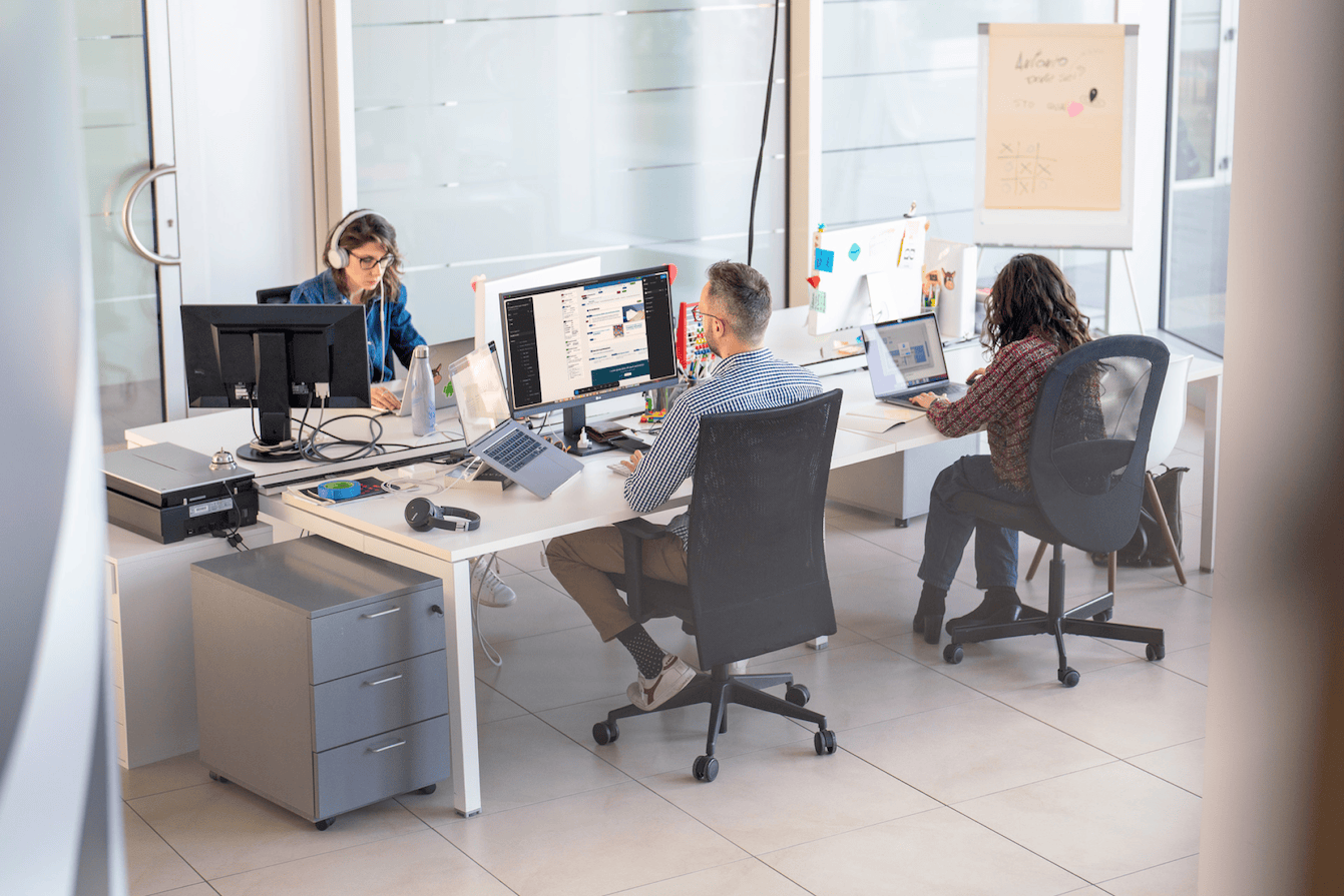 Three people sit at a shared desk, each concentrating on their computer. In the centre, one person is working on a screen with a visible digital interface. To the left, one person is wearing headphones while typing and to the right another person is working on a laptop.